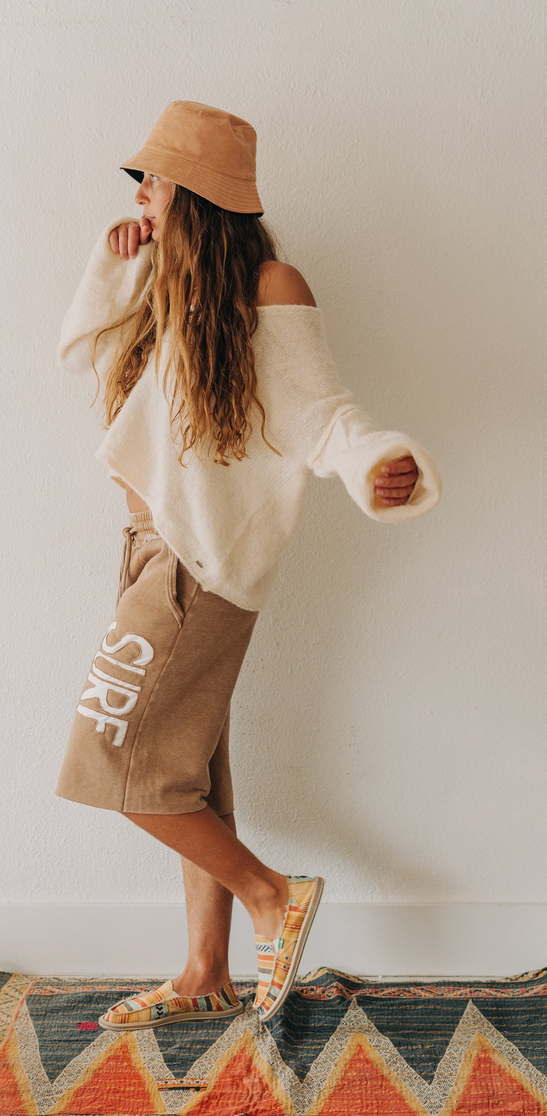 A woman wearing a beige bucket hat, a cream off-the-shoulder knit sweater, tan shorts with white lettering, and patterned sandals stands on a colorful geometric rug against a plain white wall.