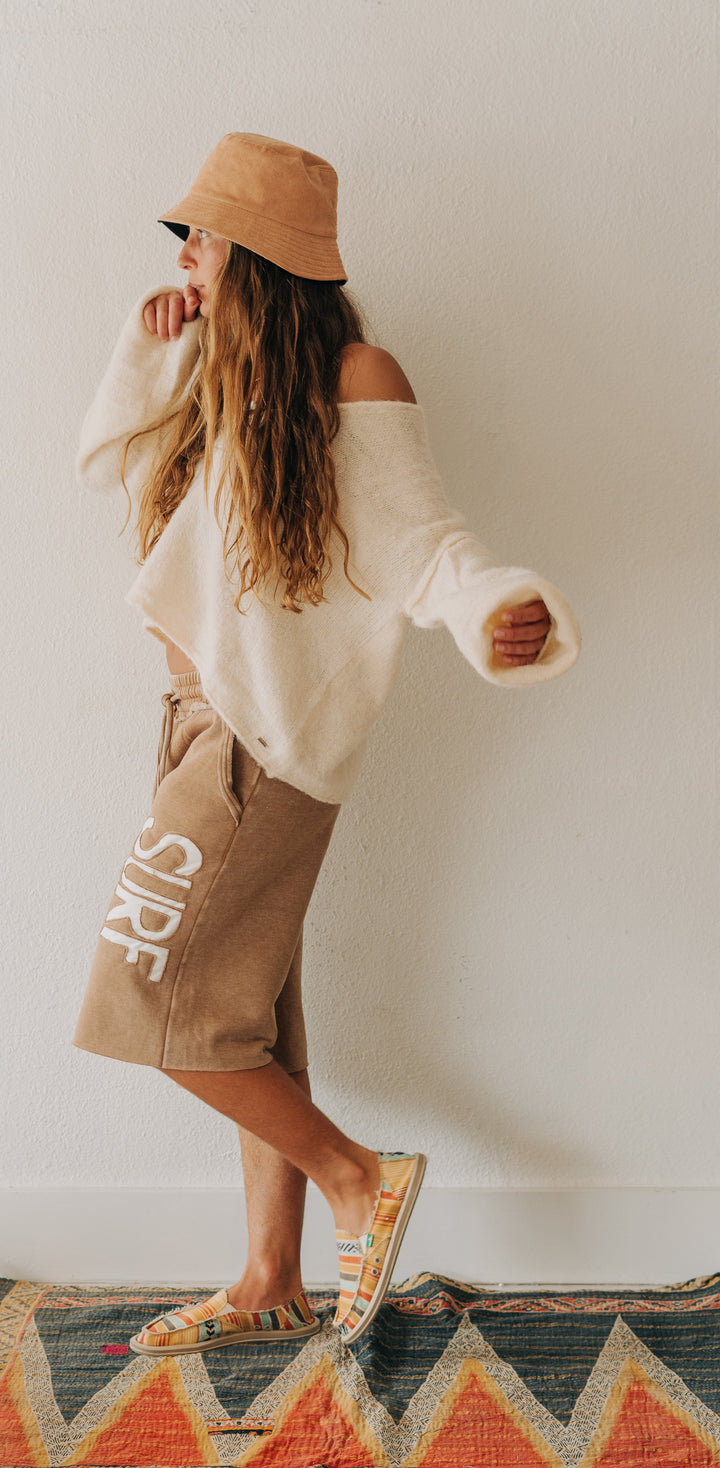 A woman wearing a beige bucket hat, a cream off-the-shoulder knit sweater, tan shorts with white lettering, and patterned sandals stands on a colorful geometric rug against a plain white wall.