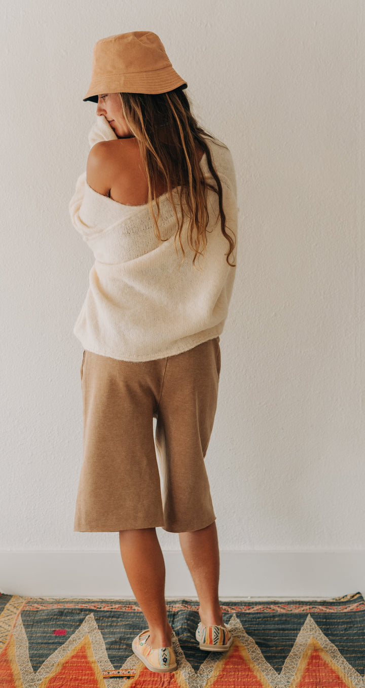 A woman stands with her back to the camera on a patterned geometric rug, wearing a tan bucket hat, a cream knit sweater draped off her shoulders, tan cropped shorts, and sandals in a bright minimalist room.