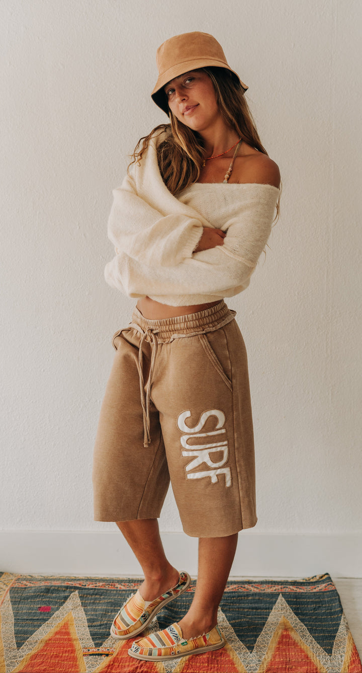 A woman stands indoors wearing tan drawstring shorts with large white &