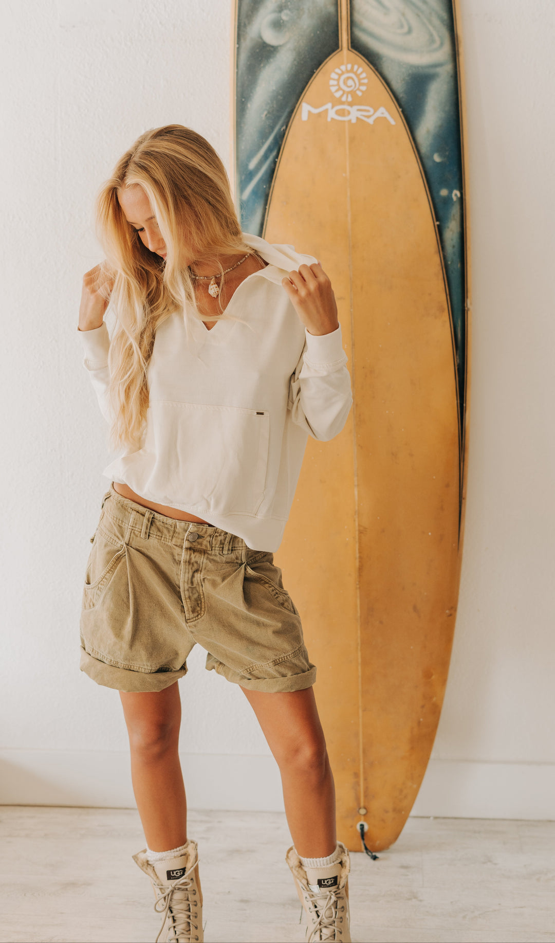A woman stands indoors wearing a white long-sleeve cropped top, beige cargo shorts, and beige lace-up boots, posing in front of a tall surfboard leaning against a white wall.