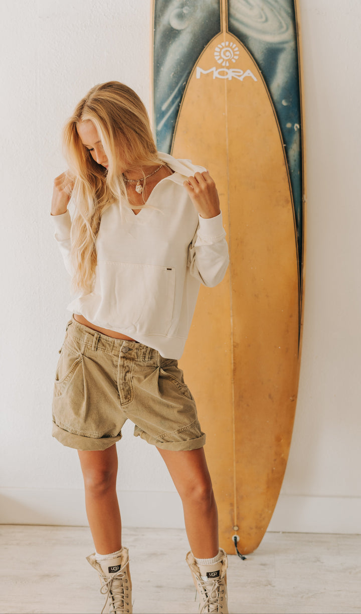 A woman stands indoors wearing a white long-sleeve cropped top, beige cargo shorts, and beige lace-up boots, posing in front of a tall surfboard leaning against a white wall.
