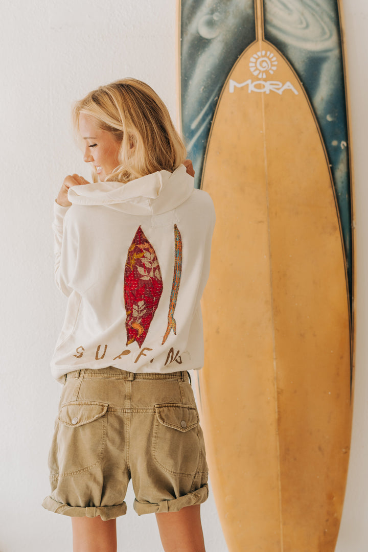 A young girl stands with her back to the camera, wearing a white hoodie with a red and gold surfboard graphic and the word &
