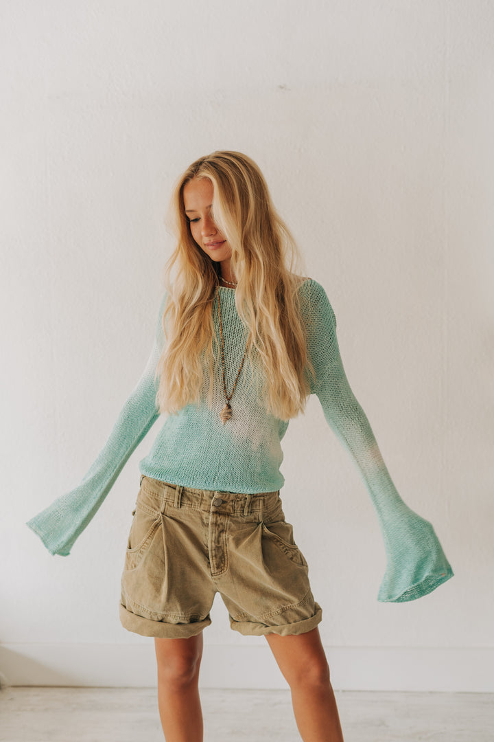 A young woman with long blonde hair wearing a fuzzy mint green long-sleeve knit sweater and khaki cargo shorts, standing against a plain white wall.
