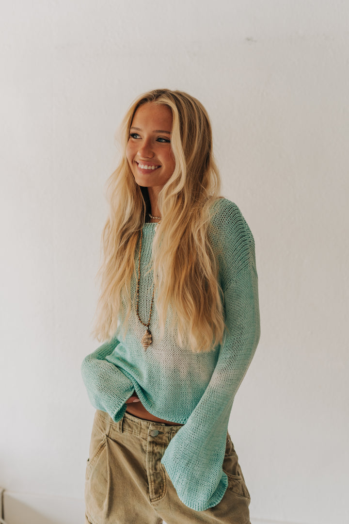 A smiling blonde woman wearing a light mint green knit sweater and khaki pants, standing against a plain white wall.