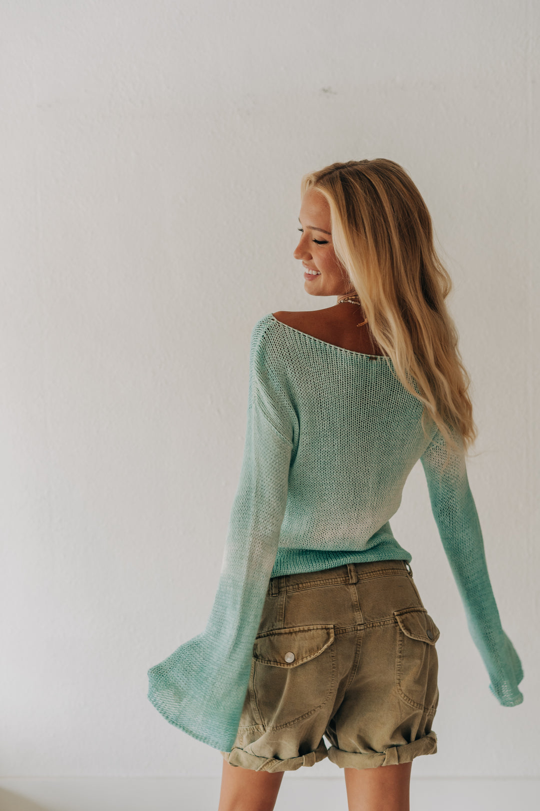 A woman with long blonde hair is shown from the back wearing a mint green knit sweater and olive green shorts against a plain white wall.