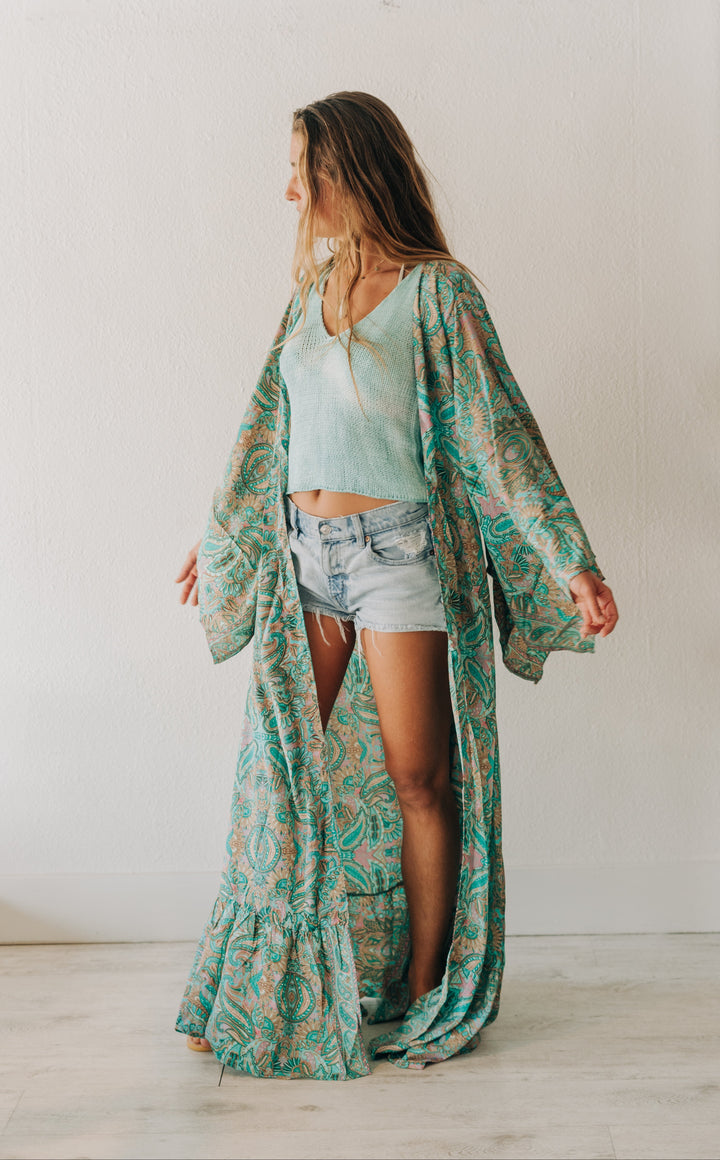 A woman stands in profile wearing a long, flowing teal and green paisley-patterned kaftan robe over a mint green crop top and denim shorts in a bright, minimalist room.