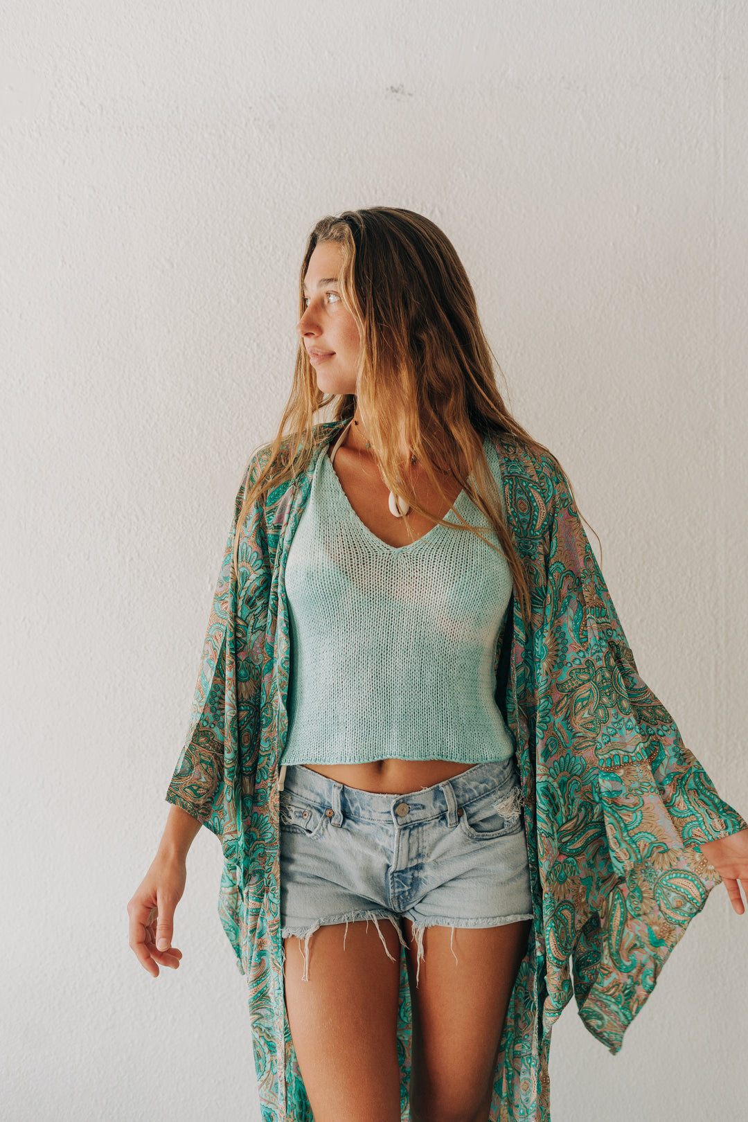 A woman wearing a light blue ribbed knit crop top, denim shorts, and a teal and brown paisley patterned open-front kimono cardigan, standing against a white wall.