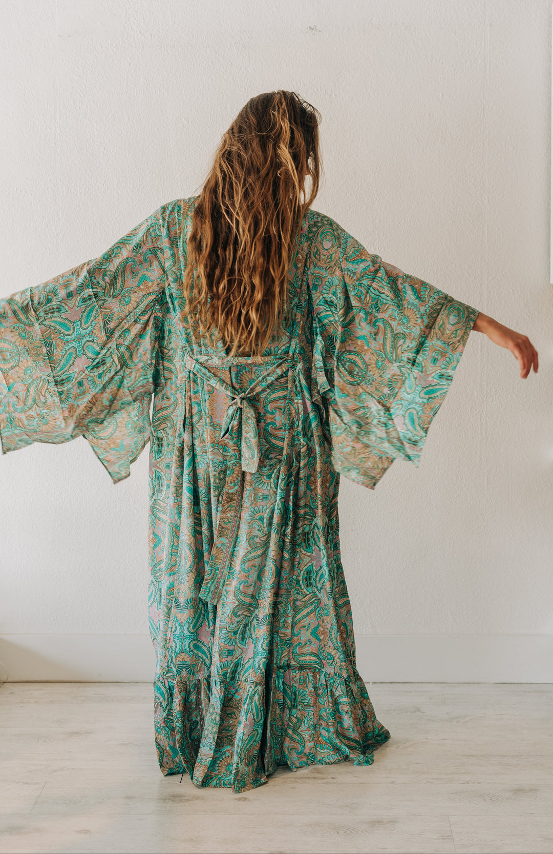 A woman viewed from behind wearing a long, flowing kaftan dress with wide bell sleeves. The dress has an intricate paisley-like pattern in teal green, brown, and beige.