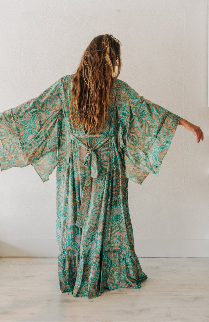 A woman viewed from behind wearing a long, flowing kaftan dress with wide bell sleeves. The dress has an intricate paisley-like pattern in teal green, brown, and beige.