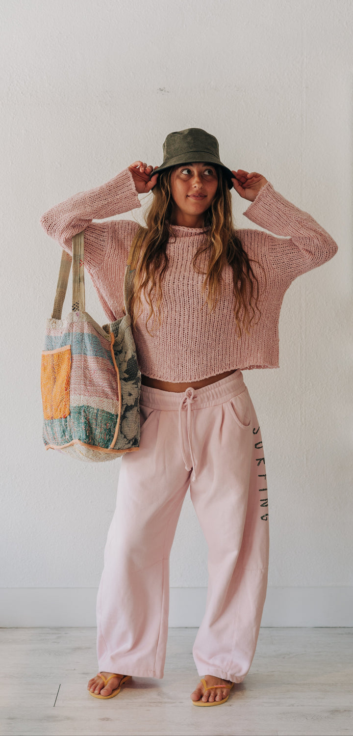 A woman stands indoors wearing a pink ribbed knit cropped sweater and matching pink sweatpants, with a green bucket hat and a multicolor patchwork tote bag. The background is a bright white room with light wood flooring and minimal shelving.