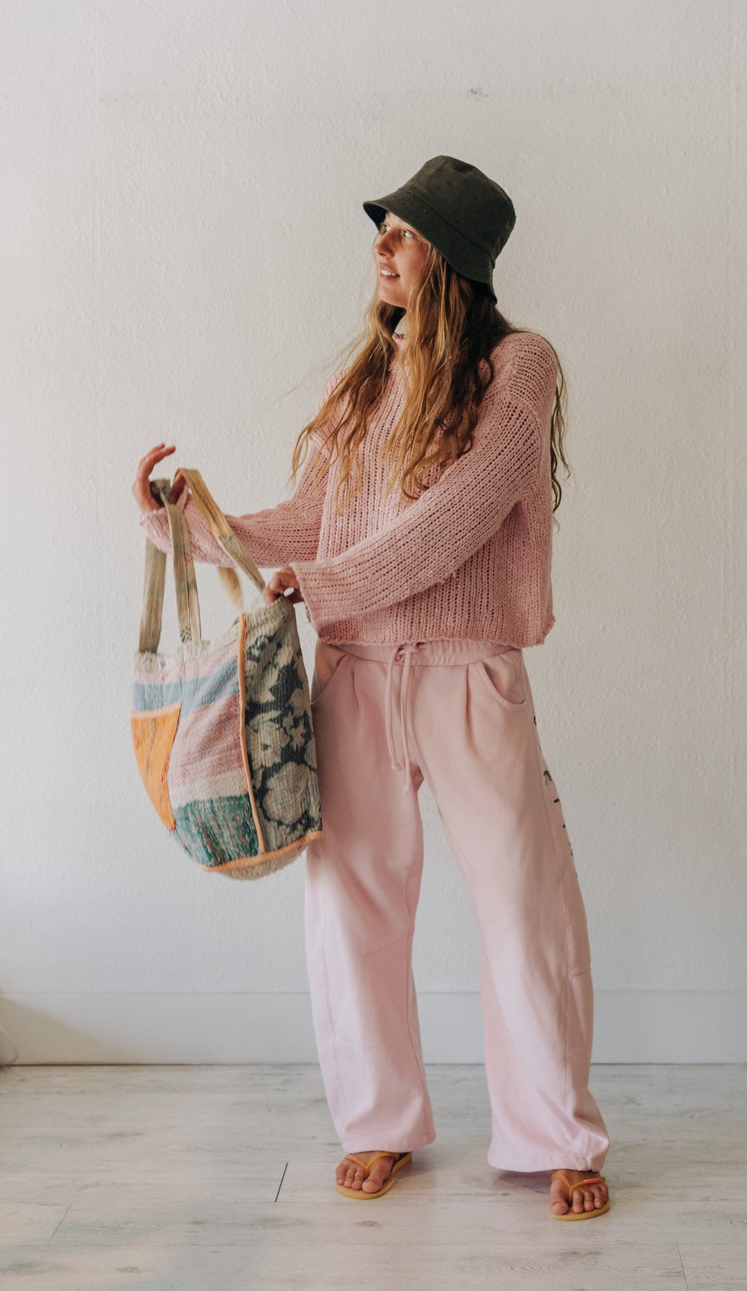 A woman stands indoors wearing a pink knit sweater and matching pink drawstring sweatpants, with a dark olive bucket hat and sandals, holding a large patchwork tote bag.