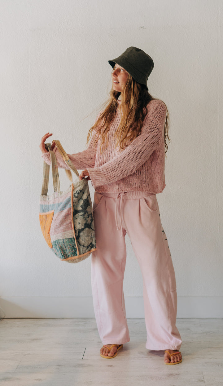 A woman stands indoors wearing a pink knit sweater and matching pink drawstring sweatpants, with a dark olive bucket hat and sandals, holding a large patchwork tote bag.
