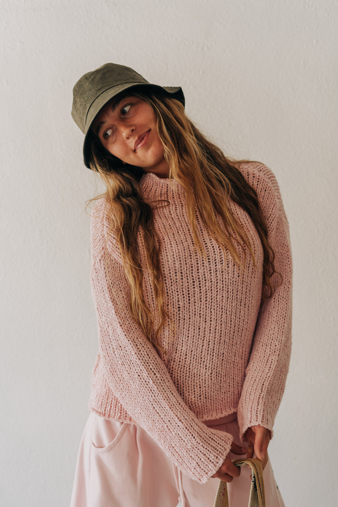 A woman wearing a chunky pink knit turtleneck sweater, light pink pants, and an olive green bucket hat, holding a beige tote bag against a plain off-white wall.