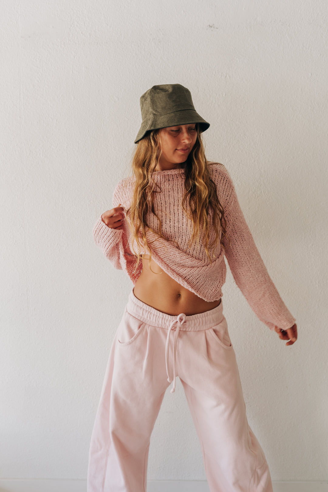 A woman modeling a pink ribbed knit cropped sweater and matching light pink sweatpants, paired with an olive green bucket hat, against a plain white wall.