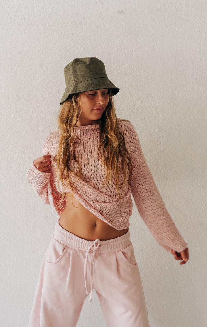 A woman modeling a pink ribbed knit cropped sweater and matching light pink sweatpants, paired with an olive green bucket hat, against a plain white wall.