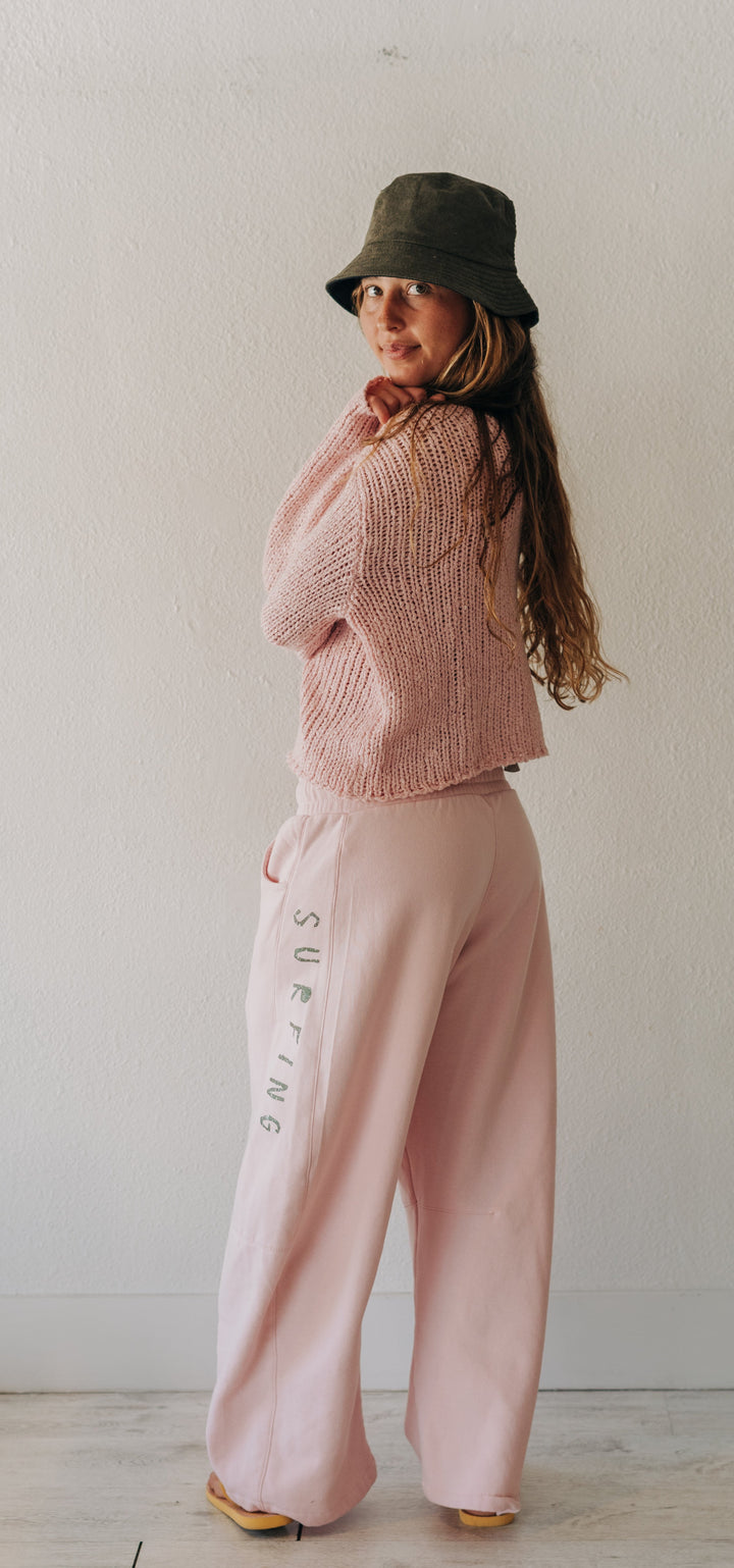 A woman stands in a minimalist room wearing a pink knit cropped sweater, matching wide-leg pink pants with vertical text, a dark olive green bucket hat, and yellow shoes.