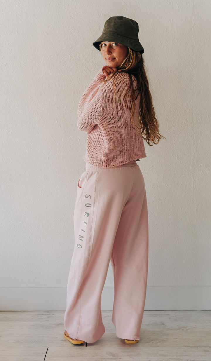 A woman stands in a minimalist room wearing a pink knit cropped sweater, matching wide-leg pink pants with vertical text, a dark olive green bucket hat, and yellow shoes.