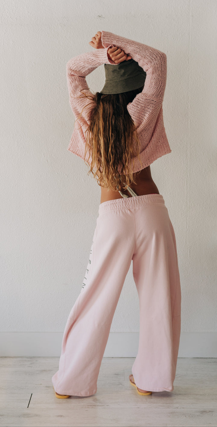 A woman stands with her back to the camera in a bright room, wearing a pink cropped knit sweater, light pink wide-leg sweatpants, and a dark olive green bucket hat.