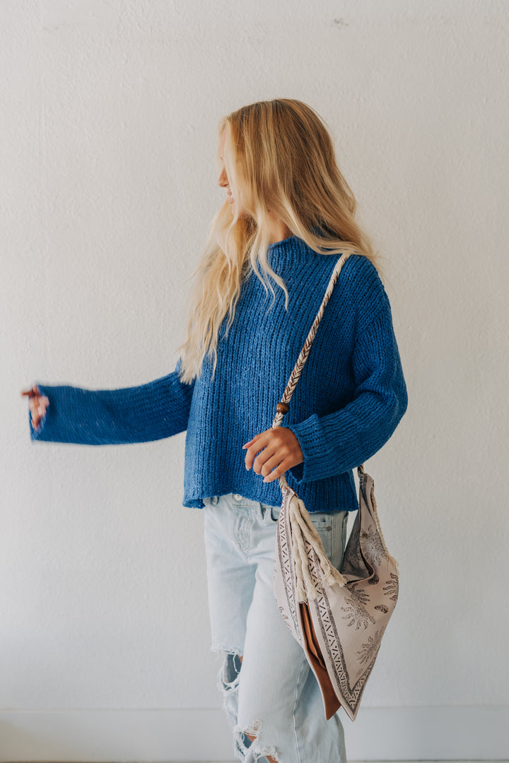 A woman with long blonde hair is shown from the side, wearing a chunky blue knit sweater, light-wash ripped jeans, and carrying a beige patterned shoulder bag against a plain white wall.