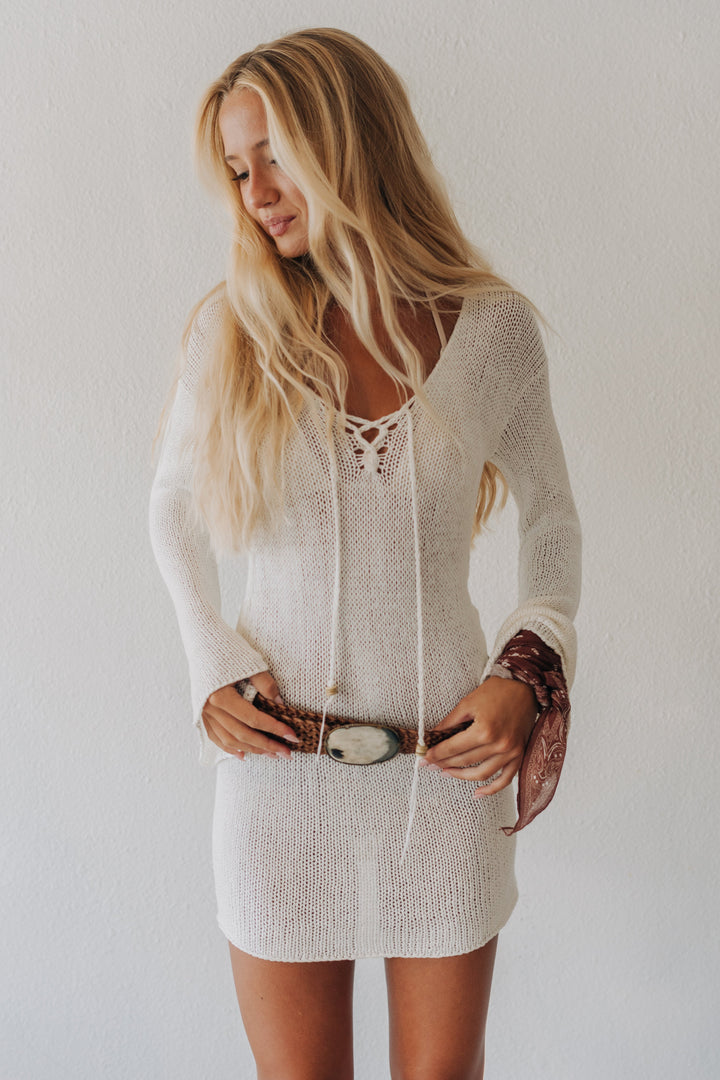 A woman with long blonde hair wearing a cream-colored, long-sleeve knit mini dress with lace-up front detailing and a matching belt, standing against a plain white wall.
