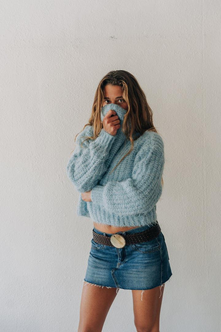 A woman wearing a chunky light blue knit sweater and a denim mini skirt with a black belt, standing against a plain white wall.