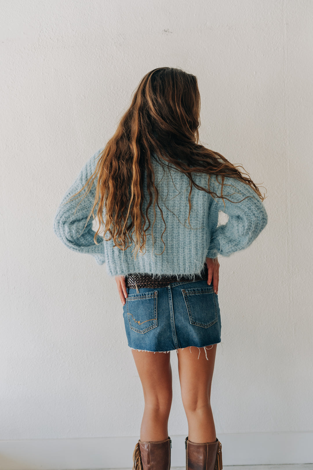 A young girl with long wavy brown hair is shown from behind, wearing a light blue knit sweater, denim shorts, and brown boots against a plain white wall.