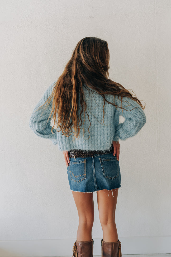 A young girl with long wavy brown hair is shown from behind, wearing a light blue knit sweater, denim shorts, and brown boots against a plain white wall.