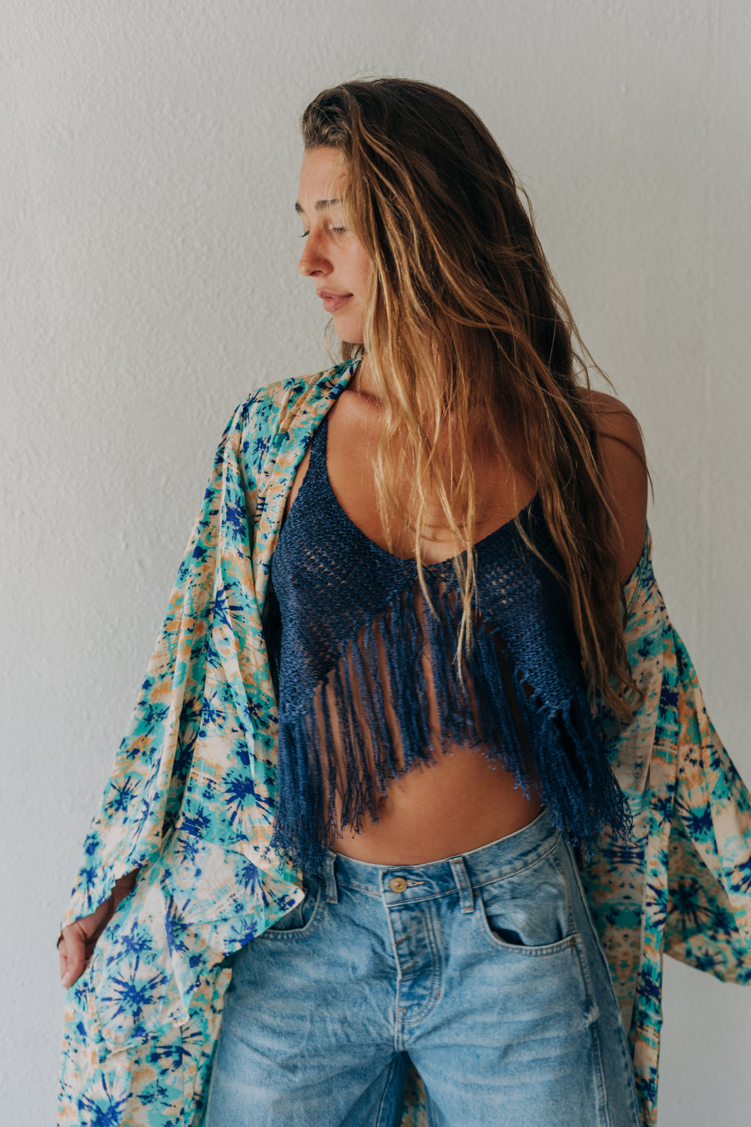 A woman wearing a dark blue fringed crochet top, light-wash blue jeans, and a colorful tropical print cardigan, posing against a neutral light gray wall.