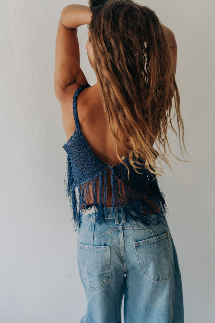 A woman with long, wavy light brown hair is shown from behind, wearing a dark blue fringed knit top and light blue denim jeans against a plain white wall.